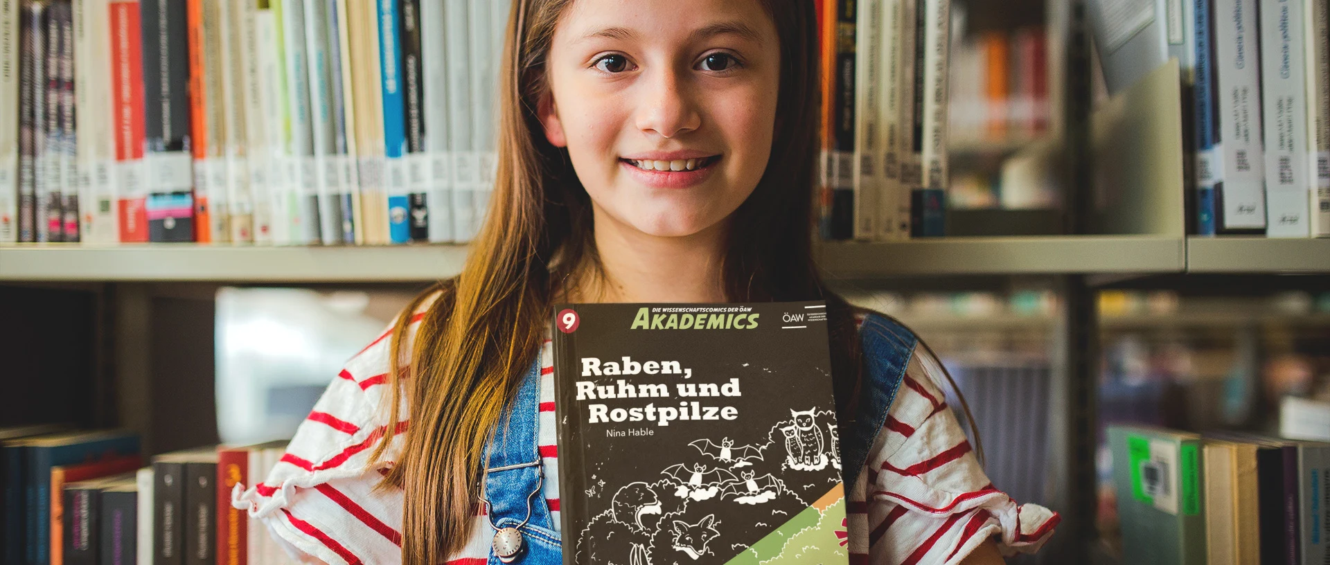 Ein Mädchen steht vor einer Bücherwand und hält eines der neuen ÖAW-Comics vor sich.